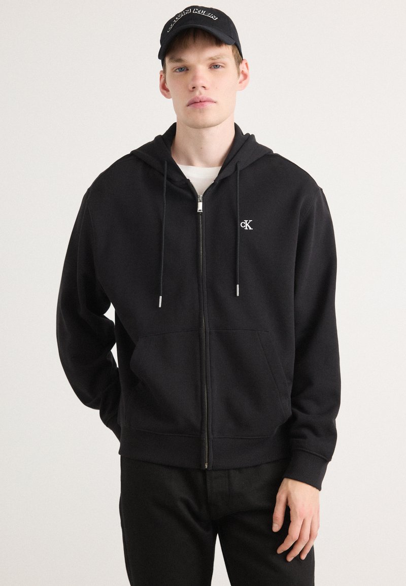 Schwarze Zip-Hoodie aus weichem Stoff. Mit einer Fronttasche, Kapuze mit Kordelzug und gesticktem Logo auf der linken Brust.