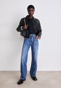 Jachetă din piele neagră cu butoane vizibile, tricou închis sub ea, blugi largi din denim albastru cu buzunare frontale și pantofi negri.
