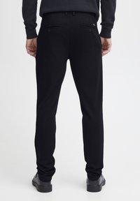 Schwarze, maßgeschneiderte Hose mit gerader Beinform aus einem glatten Stoff. Verfügt über zwei Gesäßtaschen mit Knopfverschlüssen und Gürtelschlaufen.