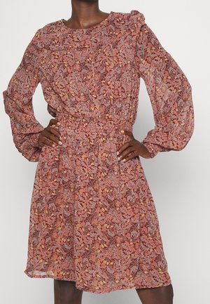 Persona de piel oscura que lleva un vestido rojo floral de manga larga con cintura ceñida y mangas abullonadas, de pie con las manos en las caderas.