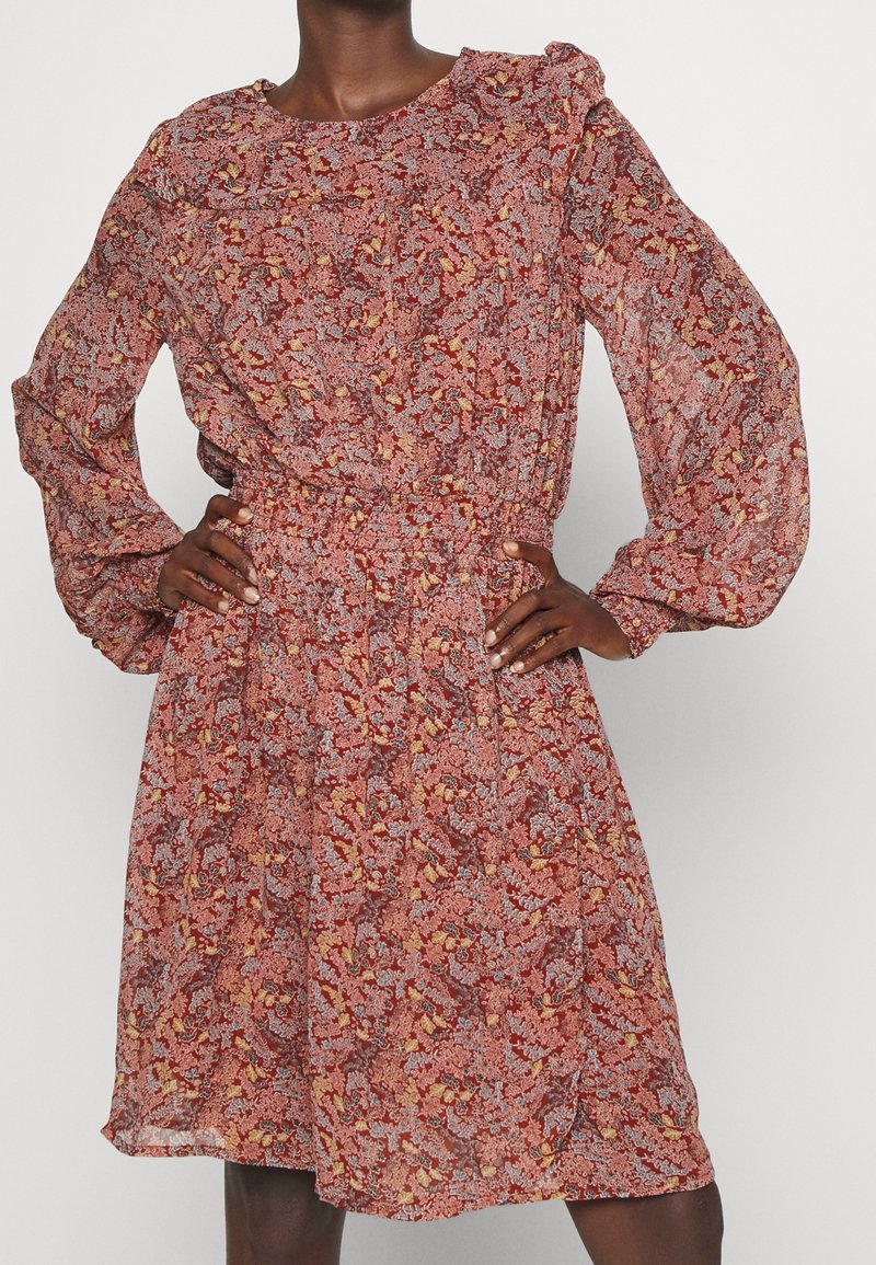 Personne à la peau foncée portant une robe rouge à fleurs avec manches longues, taille ajustée et manches bouffantes, debout les mains sur les hanches.