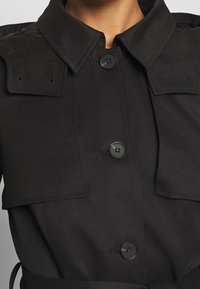 Černý trenčkot s knoflíkovým zapínáním, dvojicí náprsních kapes a límcem. Vyrobeno z hladké tkaniny se strukturovaným designem.