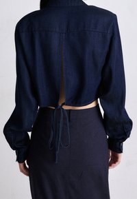 Cropped top in denim navy con maniche lunghe, con polsini a bottone e schiena scoperta legata con tessuto. Abbinato a una gonna a matita scura a vita alta.