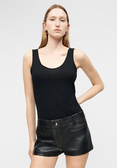 Γυναίκα με μακριά ξανθά μαλλιά που φοράει χρυσούς κρίκους στα αυτιά, μαύρο αμάνικο τοπ και μαύρο δερμάτινο σορτς, στεκόμενη με το ένα χέρι πίσω από την πλάτη.