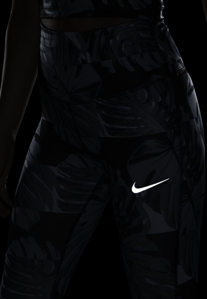 Czarne leginsy z wzorem liści, z białym logo Nike na udzie. Wykonane z gładkiego, przylegającego materiału.