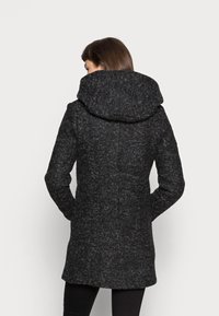 Črn teksturiran plašč z velikim kapuco, z oprijetim krojem in dolgimi rokavi. Tkanina prikazuje subtilen flekasti vzorec.