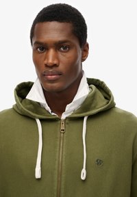 Sudadera verde con cremallera y cordón blanco, que presenta un bolsillo delantero y un pequeño logo bordado. Llevada sobre una camisa blanca con cuello.