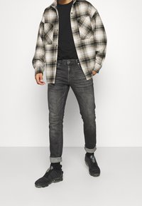 Košile z bavlněného flanelu v béžové a černé barvě, černé tričko, šedé džíny a černé tenisky s texturovaným materiálem, viditelnými švy a ohrnutými lemy.