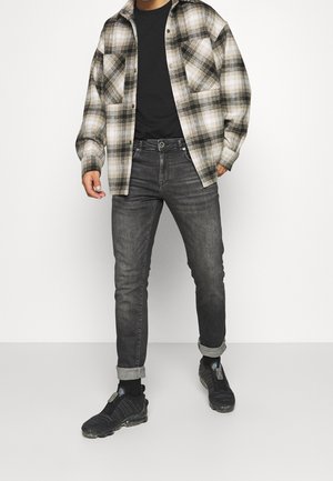 Camisa de franela a cuadros en beige y negro, camiseta negra, vaqueros grises y zapatillas negras con material texturizado, costuras visibles y puños remangados.