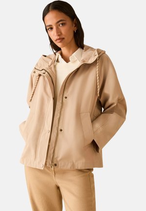 Donna che indossa una giacca beige con cappuccio sopra un maglione bianco e una camicia con colletto, con le mani nelle tasche della giacca su uno sfondo uniforme.