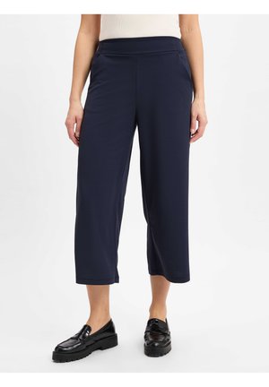 Femme portant un pantalon large cropped bleu marine et des mocassins noirs, debout devant un fond blanc uni.