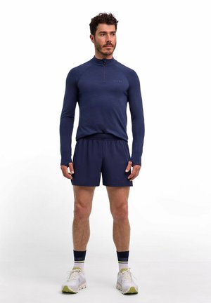 Maglia a maniche lunghe blu navy con mezza cerniera e fori per il pollice, abbinata a pantaloncini blu navy. La modella indossa scarpe da ginnastica bianche con dettagli verdi e calzini a righe.