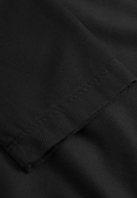 Tissu noir avec une texture lisse, présentant un ourlet replié et des coutures visibles. La surface possède un motif en côtes diagonal.
