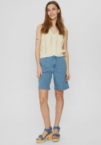 Gebreide vest in crème met een V-hals en knoopsluiting, gecombineerd met blauwe denim shorts die boven de knie eindigen, en lichtblauwe plateau sandalen.