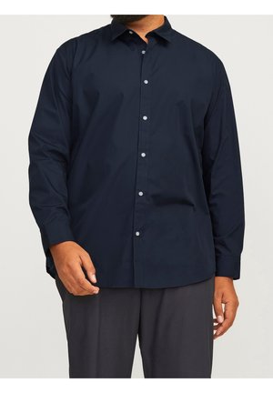 Mężczyzna w ciemnogranatowej, zapinanej na guziki koszuli z długim rękawem oraz ciemnoszarych spodniach, stojący na jednolitym jasnym tle.