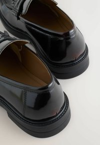 Black patent leather shoes with a sleek design, rounded toe, and contrasting brown inner lining. Features a low, textured rubber sole.