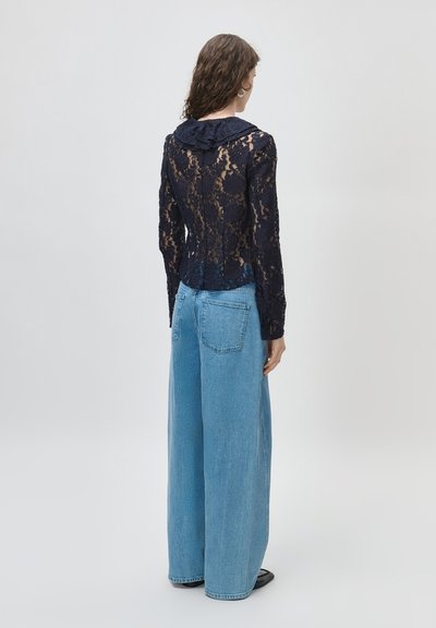 Kobieta ubrana w prześwitującą czarną koronkową bluzkę z długim rękawem, falbaniastego kołnierza oraz szerokie jasnoniebieskie dżinsy, stojąca na jednolitym tle.