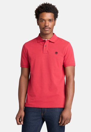 Giovane uomo con capelli corti e ricci che indossa una polo rossa a maniche corte e jeans scuri, in piedi davanti a uno sfondo neutro.