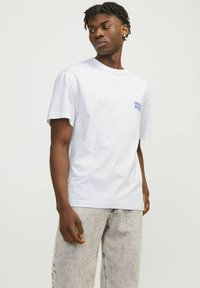Joven con rastas cortas que lleva una camiseta blanca con un pequeño texto azul y unos pantalones vaqueros claros desvanecidos, mirando de lado contra un fondo liso.