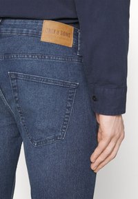 Jeans de mezclilla de color azul oscuro con un distintivo bolsillo trasero en forma triangular y una etiqueta de marca en cuero marrón con texto en relieve.