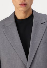Grå kavaj i strukturväv, med spetsiga slag och en enskild bröstficka, bärs över en svart t-shirt med rund halsringning. Närbild.