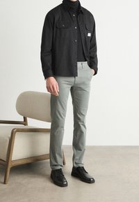 Černá košile na knoflíky s dvěma náprsními kapsami, v kombinaci se světle zelenými kalhotami a černými botami. Jednoduchý, strukturovaný design, minimální detaily.
