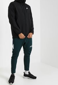 Svart lättviktsjacka från Nike med huva, dragkedjestängning och framfickor. Kombinerad med gröna smala joggers som har en vit logotyp och detaljer.