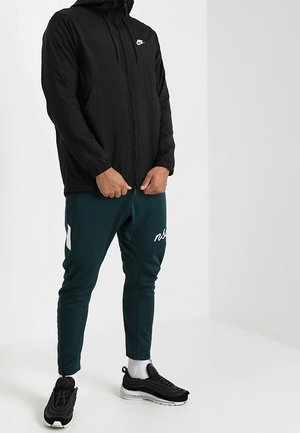 Sort letvægts Nike-jakke med hætte, lynlåslukning og frontlommer. Sammen med grønne tapered joggers med et hvidt logo og detaljer.