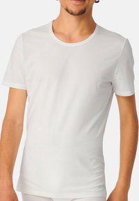 T-shirt blanc à manches courtes fabriqué en tissu léger avec une texture côtelée et un col rond. Présente un petit logo bleu au niveau de l'ourlet.