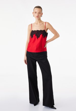 Kvinde iført rød ærmeløs top med sort blonderdetalje og sorte bukser med vide ben, stående med den ene hånd på hoften foran en ensfarvet baggrund.
