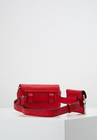 Sac banane en cuir rouge avec sangle ajustable et petite pochette attachée, présenté sur une surface blanche contre un fond clair uni.