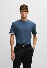 Blå bomullströja med rundad halsringning, korta ärmar med kontrasterande randiga manschetter, tillsammans med svarta byxor och ett bälte.