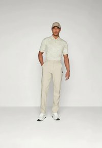 Mann med beige caps, lyse mønstrede polo skjorte, beige bukser og hvite sneakers som står med den ene hånden i lommen mot en ensfarget bakgrunn.