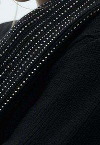 Schwarzer, strukturierter Stoff mit einem auffälligen Muster aus kleinen silbernen Nieten entlang des Ausschnitts, das ein geripptes Design zeigt.