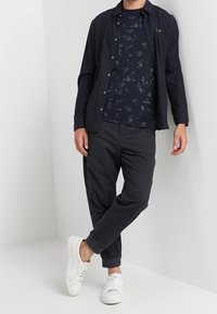 Marineblå skjorta med knappar över en mönstrad marinblå t-shirt, kombinerad med mörkgrå taperade byxor och vita sneakers, med en slät textur.