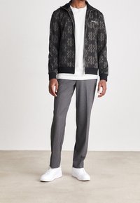 Chaqueta negra con cremallera y un elaborado patrón dorado, camiseta blanca debajo, pantalones grises y zapatillas blancas. Textura similar a la seda.
