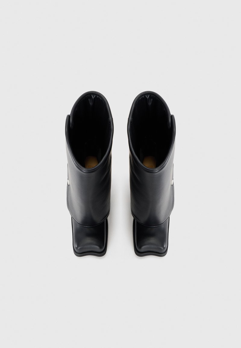 Bottes de cheville en caoutchouc noir avec un bout arrondi, texture lisse et bord replié. Présente un bouton-pression sur le côté et un design minimaliste.