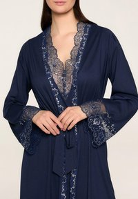 Granatowy szlafrok z dekoltem w serek, koronkowym wykończeniem, kwiecistym wzorem oraz długimi rękawami z koronkowymi akcentami. Związywany w pasie, wykonany z gładkiego materiału.
