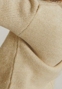 Beżowy sweter dzianinowy o miękkiej teksturze, z delikatnymi metalicznymi nićmi, które nadają blasku. Zbliżenie na detal rękawa.