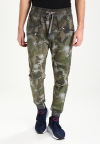 Camo-Jogginghosen in Grün und Beige, mit Reißverschlusstaschen und Kordelzugbund. Weicher Stoff mit schmalem Schnitt, kombiniert mit navyfarbenen Sneakers.