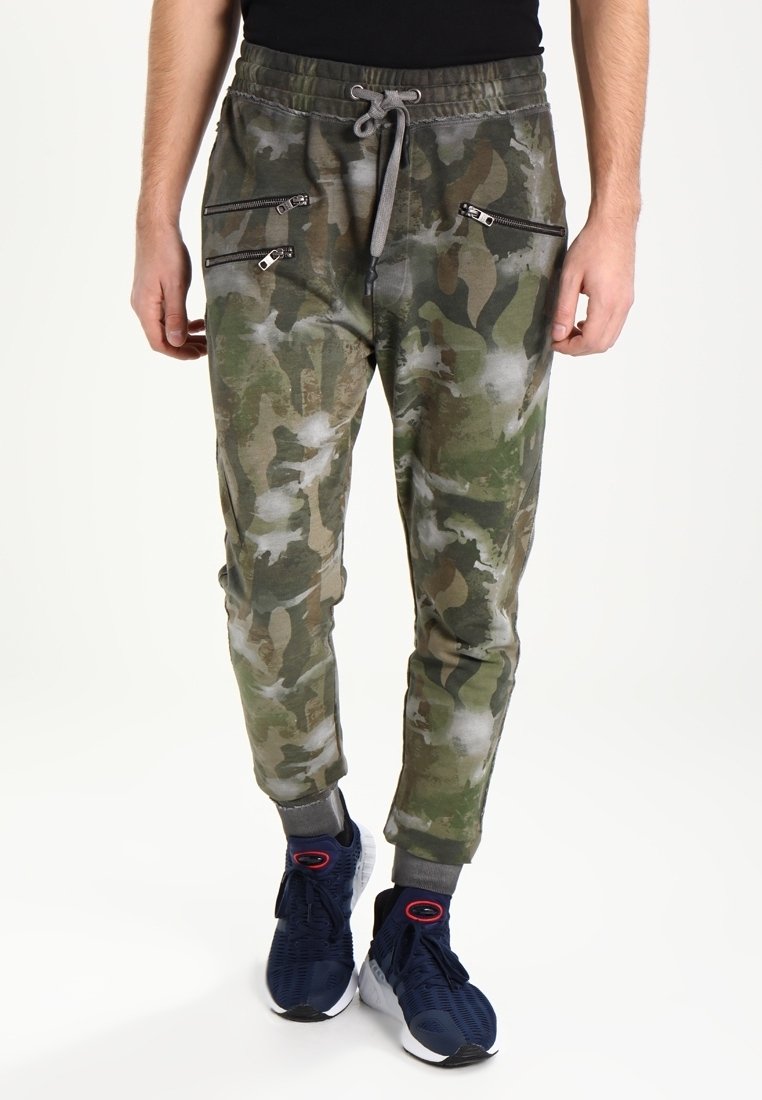 Camo-Jogginghosen in Grün und Beige, mit Reißverschlusstaschen und Kordelzugbund. Weicher Stoff mit schmalem Schnitt, kombiniert mit navyfarbenen Sneakers.