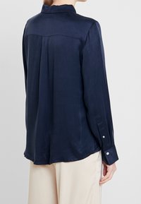 Blusa a maniche lunghe blu navy con una texture liscia, dettagli con pieghe sul retro e polsini con bottoni. Tessuto leggero e fluido che si drappeggia dolcemente.