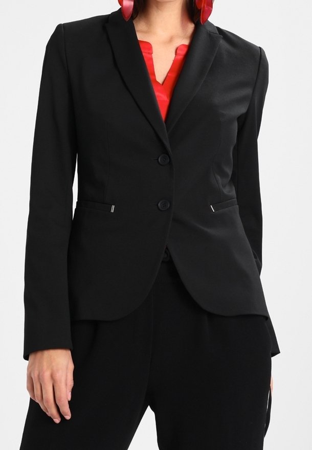 Femme portant un blazer noir ajusté sur un chemisier rouge, associé à un pantalon noir et de grandes boucles d'oreilles rouges, debout avec une main détendue.