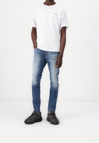 Man i vit kortärmad skjorta, blå urtvättade jeans och svarta sneakers som står med ena handen i fickan mot en enfärgad vit bakgrund.