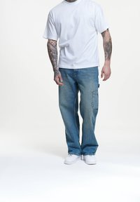 Vit t-shirt med kort ärm, ljus tvättade vida denimbukser med sidofickor och vita sneakers; tatuerade armar synliga.