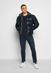 Marineblå puffjakke med en skinnende overflate, med glidelås og et mønstrete fôr. Kombinert med mørke jeans og hvite sneakers.
