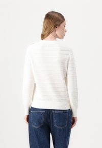 Hvit strikkegenser med horisontal ribbing, tettsittende mansjetter og rund hals, kombinert med blå høytlivede jeans.