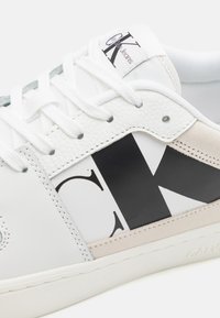 Sneaker bianco con parte superiore in pelle, dettaglio del logo nero e accenti in suede beige. Presenta lacci bianchi e una suola testurizzata.