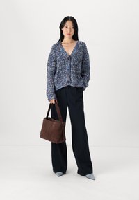 Teksturert, blå og lilla strikket cardigan med v-hals, brun semsket skinnveske og svarte vide bukser. Grå spisse sko er synlige.