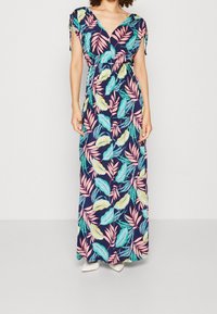 Kobieta w sukience maxi bez rękawów z dekoltem w kształcie litery V, w tropikalny motyw liści w kolorach różowym, zielonym i żółtym na granatowym tle, w połączeniu z białymi butami.